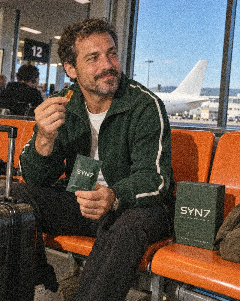 A man in a green track jacket eats a SYN7 gummy at airport gate 12, sachet in hand and the box on the seat beside him