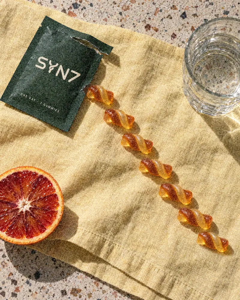 A torn SYN7 sachet and a line of seven twist gummies on a yellow linen napkin, with half a blood orange and a glass of water